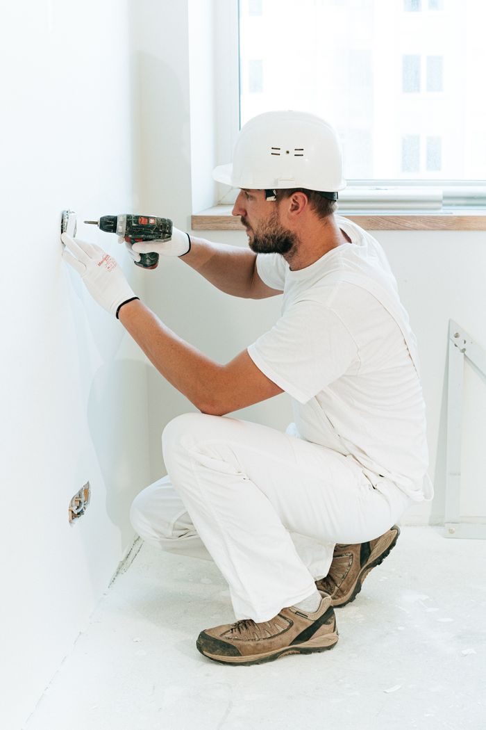 services-01 A construction worker wearing a helmet uses a drill to install a wall outlet inside an apartment.