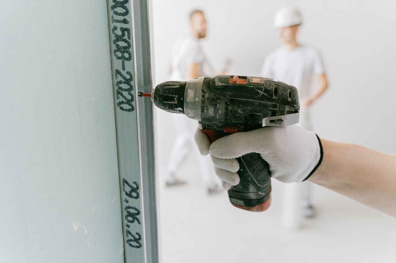 about-us-01 A construction worker uses a drill on drywall with blurred workers in the background.